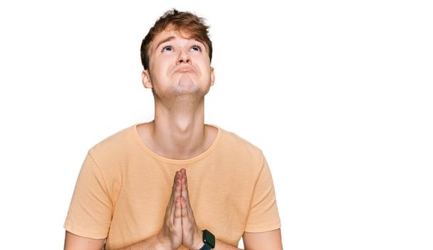 Молодий кавказький чоловік в повсякденному одязі молиться і молиться разом з надією висловлюватися на обличчі дуже емоційно і стурбовано. благання
.  - Фото, зображення