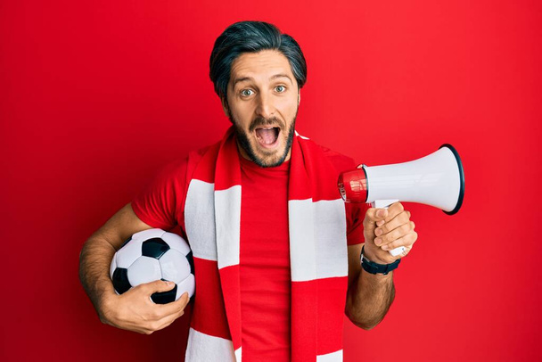 Jonge Spaanse man voetbal hooligan holding ball met behulp van megafoon vieren gek en verbaasd voor succes met open ogen schreeuwend opgewonden.  - Foto, afbeelding