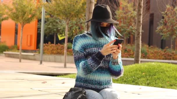 Mädchen mit blauen Haaren in schwarzer Schutzmaske und Hut mit Smartphone. Urban Fashion Outfit. - Filmmaterial, Video