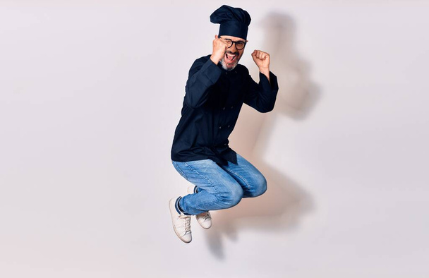 Middle age handsome hispanic man wearing glasses and cook uniform smiling happy. Jumping with smile on face celebrating with fists up over isolated white background - Foto, imagen