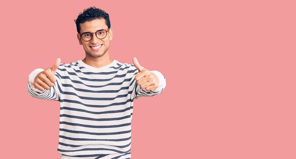Der hispanische, gut aussehende junge Mann trägt lässige Kleidung und eine Brille, die zustimmt, positive Gesten mit der Hand zu machen, Daumen hoch lächelnd und glücklich über den Erfolg. Siegergeste.  - Foto, Bild
