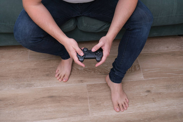 Αγνώριστος νεαρός που παίζει βιντεοπαιχνίδια με ένα gamepad που κάθεται στον καναπέ. Παίκτης στο σπίτι. - Φωτογραφία, εικόνα