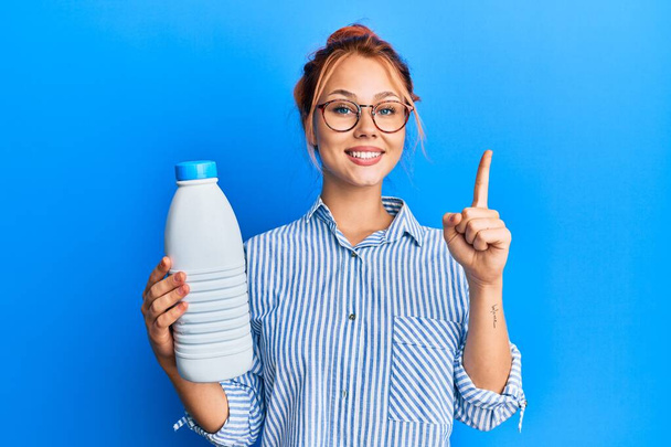Young redhead woman holding liter bottle of milk smiling with an idea or question pointing finger with happy face, number one  - Fotografie, Obrázek