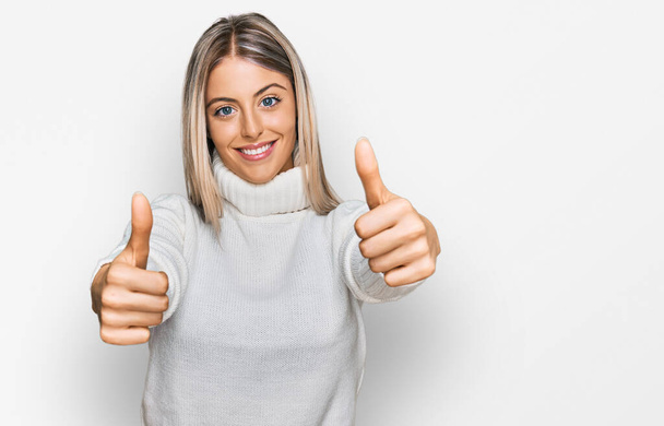 Beautiful blonde woman wearing casual turtleneck sweater approving doing positive gesture with hand, thumbs up smiling and happy for success. winner gesture.  - Photo, Image