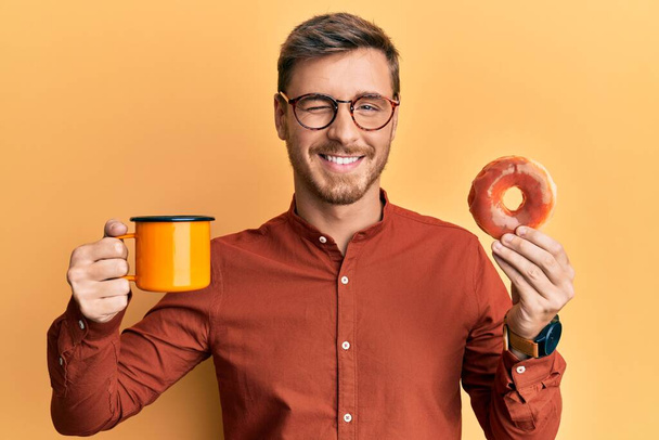 イケメンの白人男性がドーナツを食べ、コーヒーを飲みながらカメラを見てウインクしながらセクシー表情、陽気で幸せな顔.  - 写真・画像