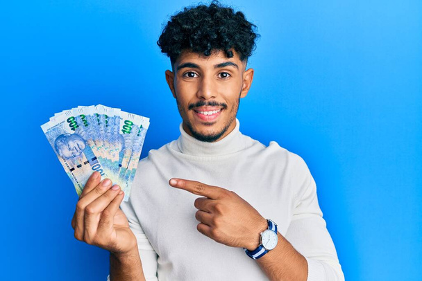 Молодий арабський красивий чоловік тримає південноафриканські банкноти 100 рандо, посміхаючись щасливо вказуючи рукою і пальцем
  - Фото, зображення