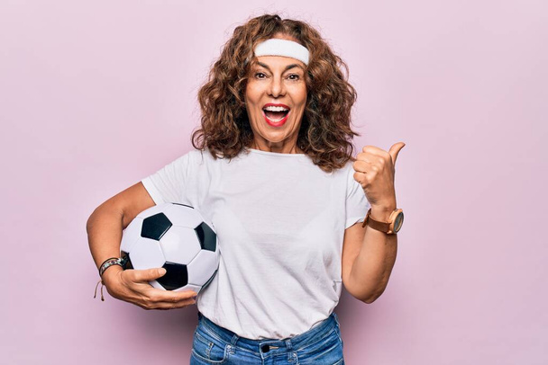 Edad media hermosa mujer deportiva jugando fútbol sosteniendo baloncesto sobre fondo rosa apuntando pulgar hacia el lado sonriendo feliz con la boca abierta - Foto, Imagen