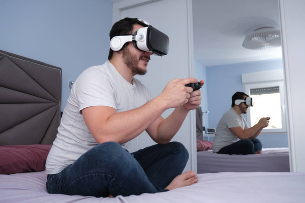 Young man playing video games with virtual reality headset and gamepad sitting on bed, reflection on wardrobe mirror. Having fun at home, video games. - Photo, Image