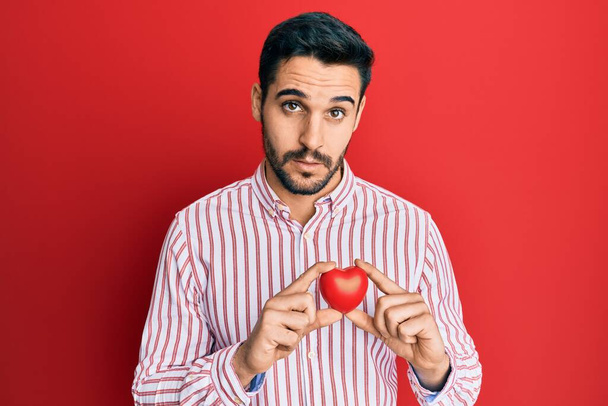 Young hispanic man holding heart clueless and confused expression. doubt concept.  - Фото, зображення