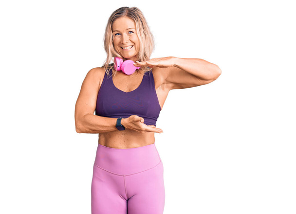 Middle age fit blonde woman wearing gym clothes and using headphones gesturing with hands showing big and large size sign, measure symbol. smiling looking at the camera. measuring concept.  - Photo, Image