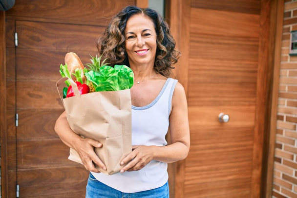 Mulher hispânica de meia idade sorrindo feliz segurando um saco de compras de supermercado cheio de mantimentos na cidade. - Foto, Imagem