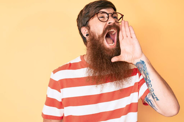 Handsome young red head man with long beard wearing casual clothes and glasses shouting and screaming loud to side with hand on mouth. communication concept.  - Photo, Image