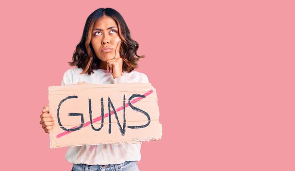 Young beautiful mixed race woman holding no guns warning banner serious face thinking about question with hand on chin, thoughtful about confusing idea  - Photo, Image