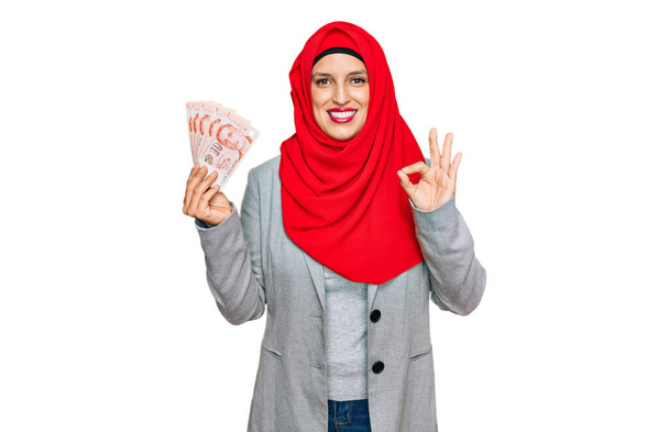 Krásná hispánská žena nosí islámský hidžáb drží singapurské dolary bankovky dělá ok znamení s prsty, usmívá přátelské gestikulace vynikající symbol  - Fotografie, Obrázek