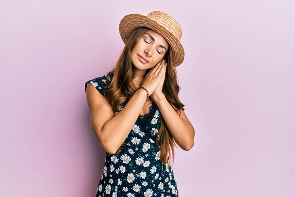 Νεαρή μελαχρινή γυναίκα που φοράει καλοκαιρινό καπέλο κοιμάται κουρασμένη ονειρεύεται και ποζάρει με τα χέρια μαζί χαμογελώντας με κλειστά μάτια.  - Φωτογραφία, εικόνα