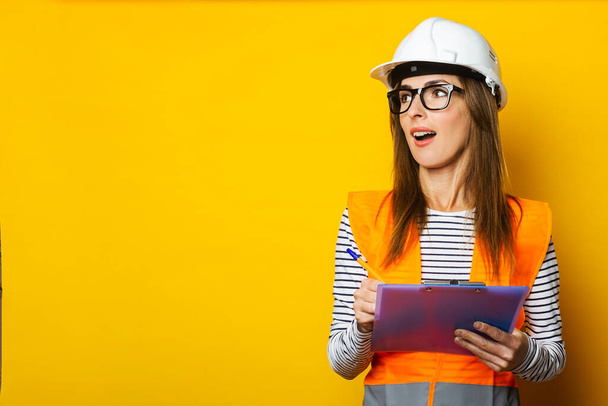 Eine junge Frau mit überraschtem Gesicht in Weste und Hut hält ein Klemmbrett auf gelbem Hintergrund. Konzept für Bau, Neubau, Sanierung. Banner. - Foto, Bild