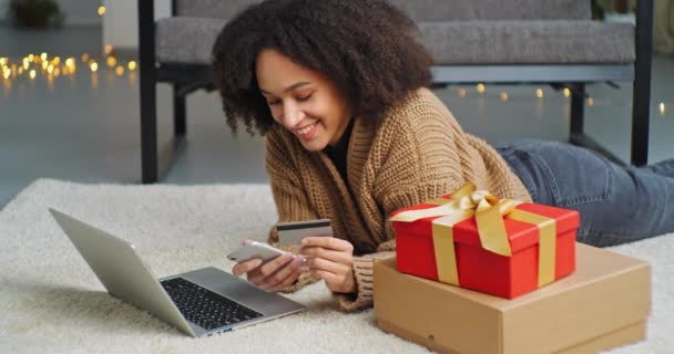 mujer afroamericana consumidor se encuentra en el suelo sobre su estómago delante de la computadora portátil cerca de la caja de regalo roja compra regalos en línea utilizando la libreta de teléfonos inteligentes móviles en las entradas de la tienda web tiene tarjeta de crédito bancaria en su mano - Imágenes, Vídeo