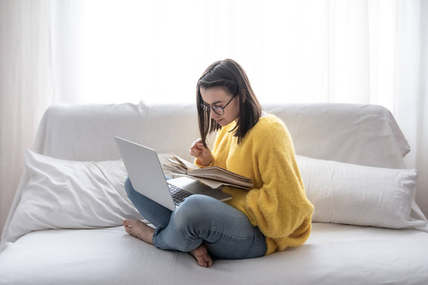Stijlvol brunette meisje in een gele trui zit thuis op de bank met een boek en laptop. Het concept van het studentenleven en zelfontwikkeling. - Foto, afbeelding