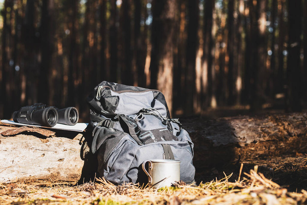 Tourist backpack, hat, binoculars and a map on a log in the forest. Concept of hiking, hiking in the mountains. Banner. - Photo, Image