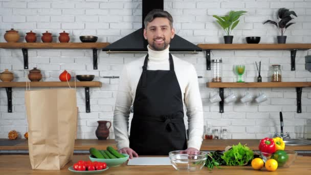 Glimlachende chef-kok in schort staat naast tafel met voedsel groenten klaar om te koken - Video