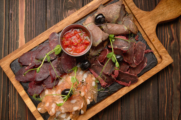 Cold cuts, sausage, basturma, smoked meat, with sauce and olives, on a wooden board, on a wooden background - Photo, Image