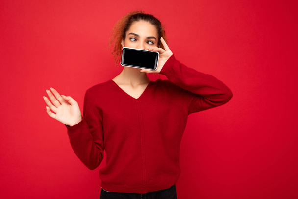 Photo of happy funny young female person wearing dark red sweater isolated over red background holding smartphone and showing mobile phone screen with copy space for cutout - Fotó, kép