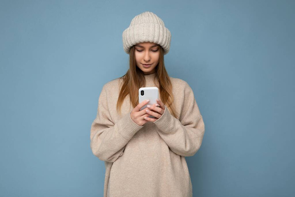 Gündelik kıyafetler giyen güzel genç bir kadın arka planda izole bir şekilde duruyor telefonla internette geziniyor ve mobil ekrana bakıyor. - Fotoğraf, Görsel