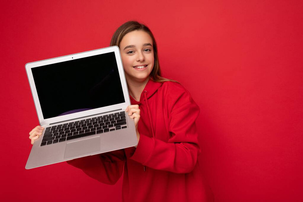 Close-up portrait photo of beautiful happy smiling girl with long hair wearing red hoodie holding computer laptop looking at camera isolated over red wall background - Zdjęcie, obraz