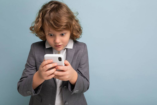 Zdjęcie Przystojnego, zszokowanego chłopca z kręconymi włosami w szarym garniturze, trzymającego i korzystającego z telefonu odizolowanego na niebieskim tle, patrzącego na smartfona, komunikującego sms-y. - Zdjęcie, obraz