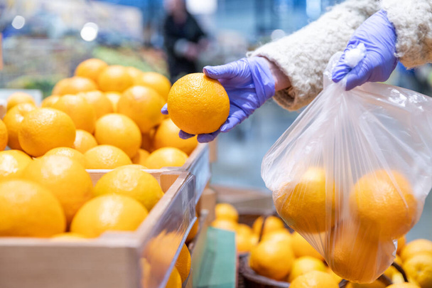 As mãos de mulher em luvas médicas escolhem laranjas frescas que se dobram em um saco plástico descartável no supermercado, perto. Medidas de protecção contra a pandemia do coronavírus, surto de covid-19. Foco seletivo.  - Foto, Imagem