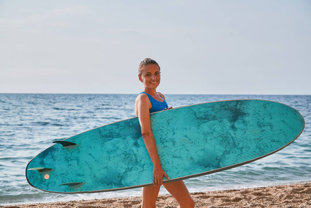 Smiling young lady carrying surfboard from ocean. Learning surfing, summer vacation and adventure time on beach concept - Fotografie, Obrázek