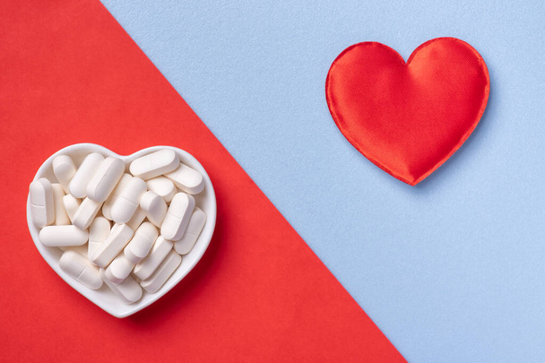 Pills in bowl and red heart on blue and red background. Medicines for cardiology health treatment and diseases. - Фото, зображення