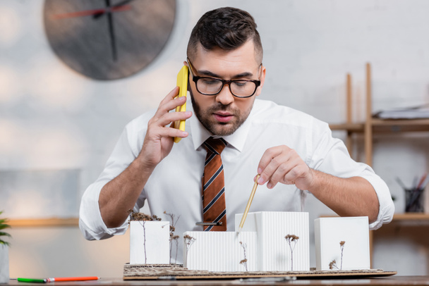 architekt ukazuje na domy modely s tužkou při mluvení na smartphonu na pracovišti - Fotografie, Obrázek