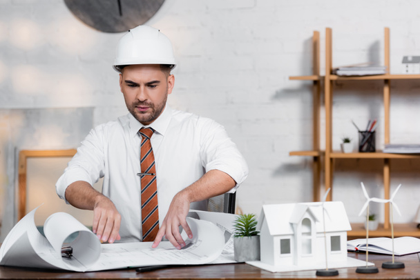 серьезный архитектор в шлеме смотрит на чертеж рядом с моделью дома - Фото, изображение