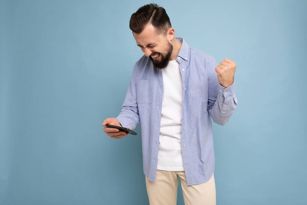 Photo shot of sad handsome bearded good looking young man wearing casual stylish outfit poising isolated on background with empty space holding in hand and using mobile phone messaging sms looking at - Photo, Image