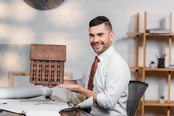 happy architect smiling at camera while pointing at house model at workplace - Photo, Image