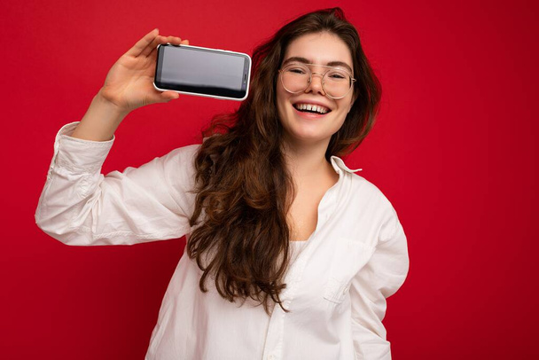 Fotografie krásné usmívající se mladá žena dobře vypadající na sobě ležérní stylové oblečení stojící izolované na pozadí s kopírovacím prostorem drží smartphone ukazující telefon v ruce s prázdným displejem obrazovky pro - Fotografie, Obrázek