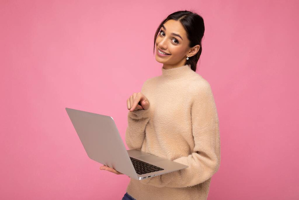Photo of Beautiful young brunette woman wearing beige sweater holding netbook computer looking at camera showing at laptop isolated over pink wall background - Fotó, kép