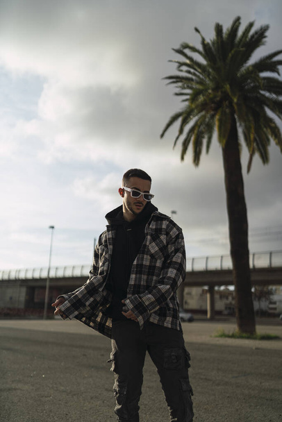 A vertical shot of a stylish guy wearing a plaid jacket, a black hoodie, black pants, and sunglasses - 写真・画像
