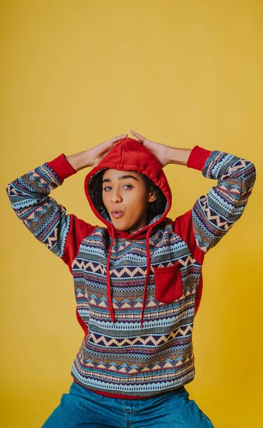 A portrait of a cheerful Hispanic female with curly hair wearing a hoodie and jeans against a yellow background - 写真・画像