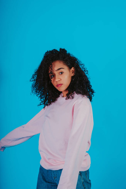 A beautiful curly haired girl posing in a pink sweater and jeans on a blue background - Valokuva, kuva