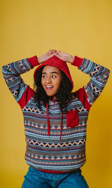 A portrait of a cheerful Hispanic female with curly hair wearing a hoodie and jeans against a yellow background - Foto, imagen
