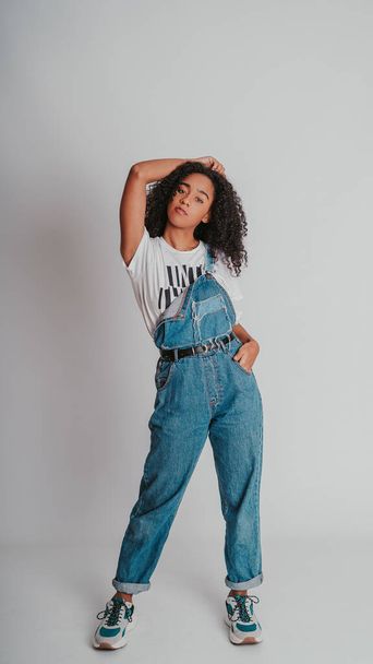 A portrait of a curly Hispanic female wearing denim overalls and posing against a wall - Zdjęcie, obraz