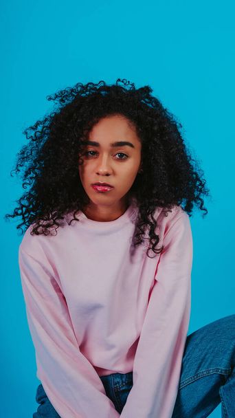 A beautiful curly haired girl posing in a pink sweater and jeans on a blue background - Foto, imagen