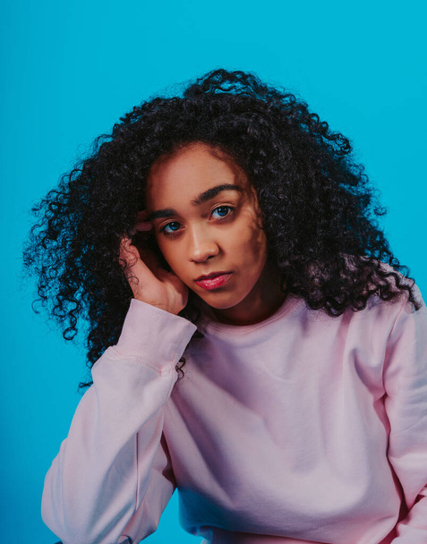 A portrait of a young Hispanic female with curly hair sitting against a blue background - Foto, Imagem