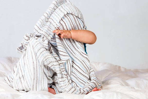 A shot of a funny little baby sitting on a bed and trying to put off a blanket from his face - Foto, imagen
