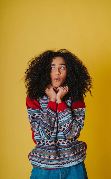 A cheerful Hispanic female with curly hair wearing jeans and a hoodie and posing against a yellow background - Photo, image