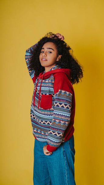 A confident young Hispanic female wearing a hoodie with jeans and posing against a yellow background - Foto, Imagem