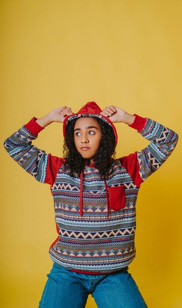 A portrait of a young Hispanic female with curly hair wearing a hoodie and jeans against a yellow background - Photo, image