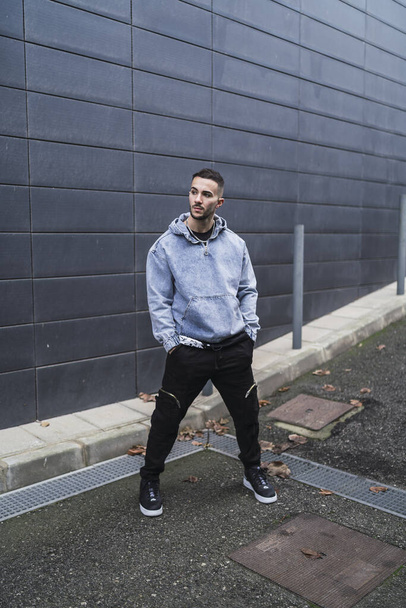 A vertical shot of a stylish guy wearing a necklace, a blue hoodie, black pants, and sneakers - Zdjęcie, obraz